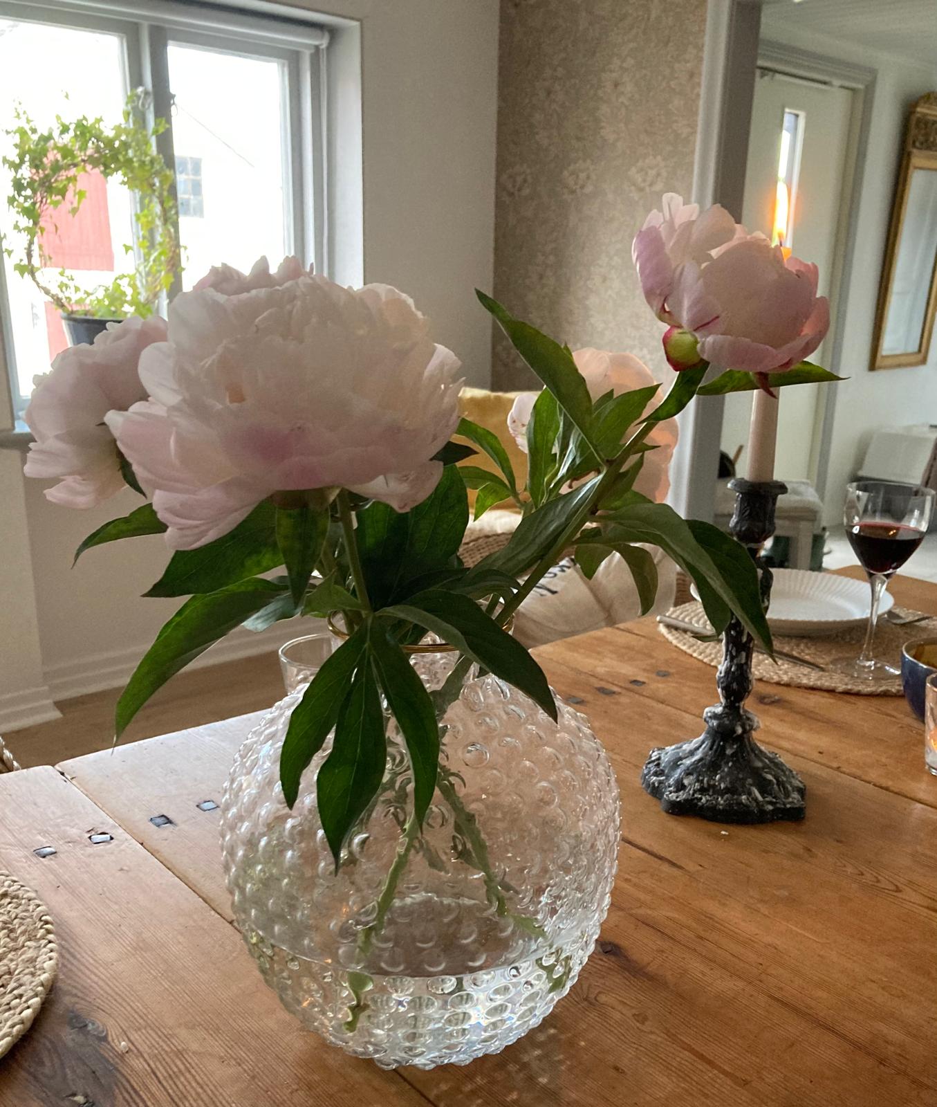 Ljusrosa pioner i en bubblig glasvas på ett dukat furubord